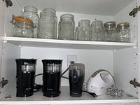 Overall view of Ball glass jars on upper shelf, two Mr. Coffee grinders, Hamilton Beach grinder, and Sunbeam hand mixer on lower shelf within cabinet.