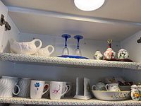 Two shelves of ceramic kitchen items arranged on floral paper-lined shelves, showing coffee cups, ceramic gravy boats, blue stemmed glasses, and condiment containers.