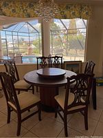 Six wooden scrolled back dining chairs with cream fabric seats around a round wooden table, viewed from a distance near bright window with pool outside.