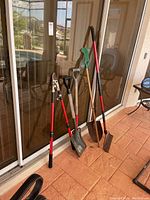 Garden tools including two shovels, pitchfork, rake, and pruner lined up against sliding door on terracotta tile floor.