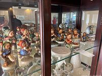 Glass display shelf with 15 painted Goebel figurines showing children in various poses, some with instruments, wreath, or baskets. Also visible are white porcelain items on lower shelf.