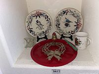 Photo of three decorative plates (two bird-themed round plates, one red holiday tray plate with wreath design), holiday-themed mug, and small glass sugar bowl.