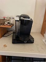 Black and silver Keurig coffee maker on counter, unplugged, with power cord shown.