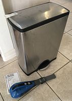 Metal trash can with pedal lid and Black & Decker cordless hand vacuum on the floor with manuals visible.
