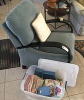 Side view of blue microfiber recliner with black wood arms and a decorative pillow, plastic tub with assorted linens in foreground.