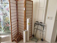 Three-panel wooden room divider with translucent panels folded, placed near a doorway with a floral curtain in the background. Rattan magazine holder and rolled posters are placed at the base. Walker positioned to the right.