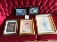 Five framed art pieces displayed on a red sofa, showing variety in subjects and frame styles.