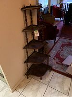Wooden five-tier corner shelf unit seen from front left side against a wall corner, showing its shape, turned supports, and size.