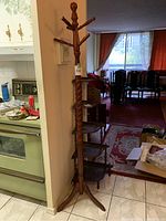 Full view of the hardwood jacket rack standing in a tiled kitchen entryway, showing its multiple pegs and tripod base with additional pegs included.