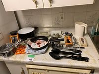 Overview of the lot showing all items on a kitchen counter including pots, pans, utensils, scale, cookie cutters, and packaged kitchen gadgets.