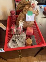 Overall view of a red plastic basket containing various unused candles: bagged white tea lights, two angel-shaped candles in boxes with red ribbons, a large red pillar candle, a gold votive candle, and some smaller votive candles.