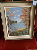 Framed landscape painting showing autumn trees, water, rocks, and sky.