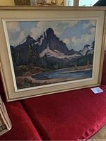 Framed landscape painting showing mountains, lake, and trees on a red surface.