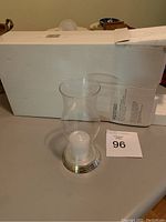 Single glass hurricane candle holder with silver plated base positioned next to white box.