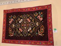 Black tapestry with pink and white embroidered elephants, floral and leaf designs, with a red and orange border hung on a wall.