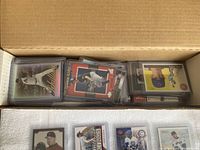 Box containing approximately 40 baseball cards in plastic top loaders, showing a variety of player cards stacked inside a cardboard box.