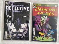 Two comic books side by side in protective bags, one showing Batman's face and the other showing Joker with guns.