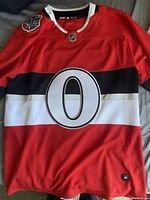 Front view of red Ottawa Senators hockey jersey with large 'O' logo and horizontal black and white stripes across the chest.