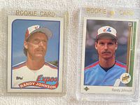 Two Randy Johnson rookie baseball cards displayed side by side. Left card is 1989 Topps #647 in protective top loader, right card is 1989 Upper Deck #25 in magnetic rookie card holder.