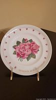 Photo shows a white round ceramic plate with a central design of two large pink roses with green leaves and a border featuring small pink rosebuds. The text 'Fifteenth 40th Anniversary' is printed near the edge in gold color. The plate is displayed on a stand against a plain background.