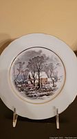 Front view of the decorative plate showing the winter log cabin scene with gold trim and scalloped edges.