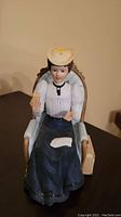 Photo shows porcelain figurine of nurse Mrs. Albee seated in detailed chair wearing white nurse cap and dark skirt, holding glass and towel.