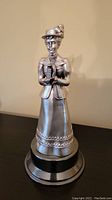Metal figurine trophy shaped as a woman in period costume holding a trophy, on round reflective base.