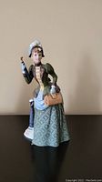 Porcelain figurine of a woman in green patterned suit dress holding handbag and umbrella against a beige wall