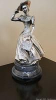 Silver-colored metal figurine of a woman in Victorian dress holding a handbag and tipping her hat, mounted on a black base labeled 'DISTRICT AWARD'.
