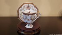 Porcelain teacup and saucer with portrait and floral garlands shown on display stand.