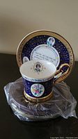 Tea cup and saucer set featuring blue background with gold accents and floral and portrait images, placed on a clear plastic covering.