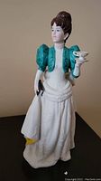 Porcelain figurine of a woman wearing a white gown with green shrug and holding tea cup, shown against a neutral background.
