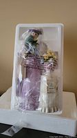 Photo showing a figurine of a woman in a lavender skirt with flowers, secured in clear styrofoam packaging along with a white pedestal with plaque.