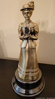 Front view of the bronze-colored female figurine dressed in period attire holding binoculars, mounted on a black round base.