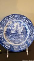Blue and white ceramic plate featuring an illustration of Independence Hall at the center, with decorative border including leaves and doves.