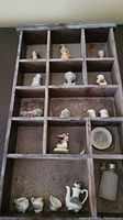 Wooden display wall case divided into small compartments with various Avon porcelain miniature figurines, teapot set, and bottle inside. Case shows signs of wear and dust.