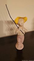 Porcelain figurine of a boy wearing a yellow hat holding a fishing pole, standing on a black surface against a plain background