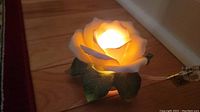 Porcelain night light shaped like a white rose with green leaves, glowing warmly on wooden surface near baseboard.