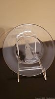 Image shows a clear glass round plate with an embossed figure of a woman standing by a swing or frame structure.