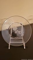 Clear round glass plate standing on a display stand. Central embossed rose floral design visible.