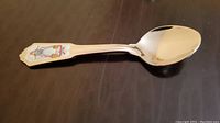 Photo of a silver-toned souvenir teaspoon with a decorative handle showing a colorful emblem of a German holiday scene or figure.