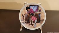 Round decorative plate with a painted image of a mother and two children near a fireplace and mirror, displayed upright on a stand.
