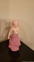 Figurine of a woman standing on a flat surface, wearing a pink dress and shawl, missing a hoop attachment.