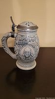 Photo showing a ceramic beer stein with embossed postal service themed decorations including a portrait of Benjamin Franklin under the lid. Pewter lid with broken decorative tip remains attached and can close.