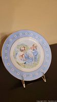 Front view of commemorative plate with scene of woman holding baby and child nearby, blue floral border