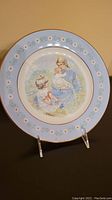 Photo of commemorative decorative plate showing a woman holding a baby with a girl beside them, light blue rim with white daisy pattern and gold trim