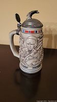 Front view of ceramic beer stein with detailed relief of American Armed Forces personnel, colored ribbons, and pewter lid with eagle finial