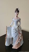 Porcelain figurine depicting a woman in white and pink gown holding book and small dog, next to wooden chair with draped white fabric.
