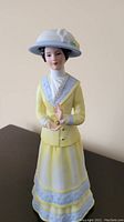 Front view of porcelain figurine of woman in yellow dress holding perfume bottle on black surface with neutral background.