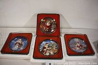Four square collector tins in red and black with corresponding ceramic plates inside, each featuring Christmas-themed artwork.